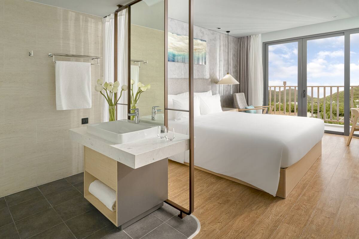 A modern hotel room with a large bed, a bathroom with a sink and mirror, and a balcony with a view.