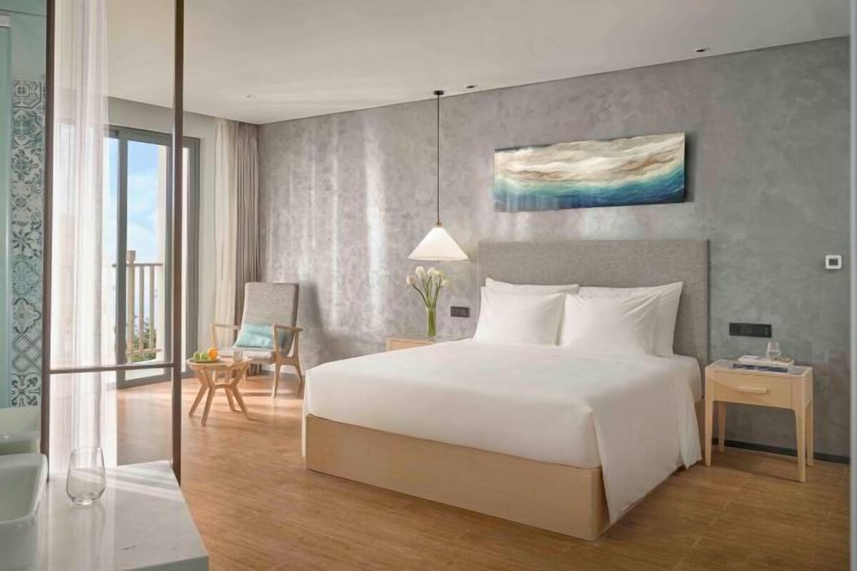Una habitación de hotel con una cama grande, una mesita de noche, una silla, un cuadro en la pared y vistas a través de una ventana.