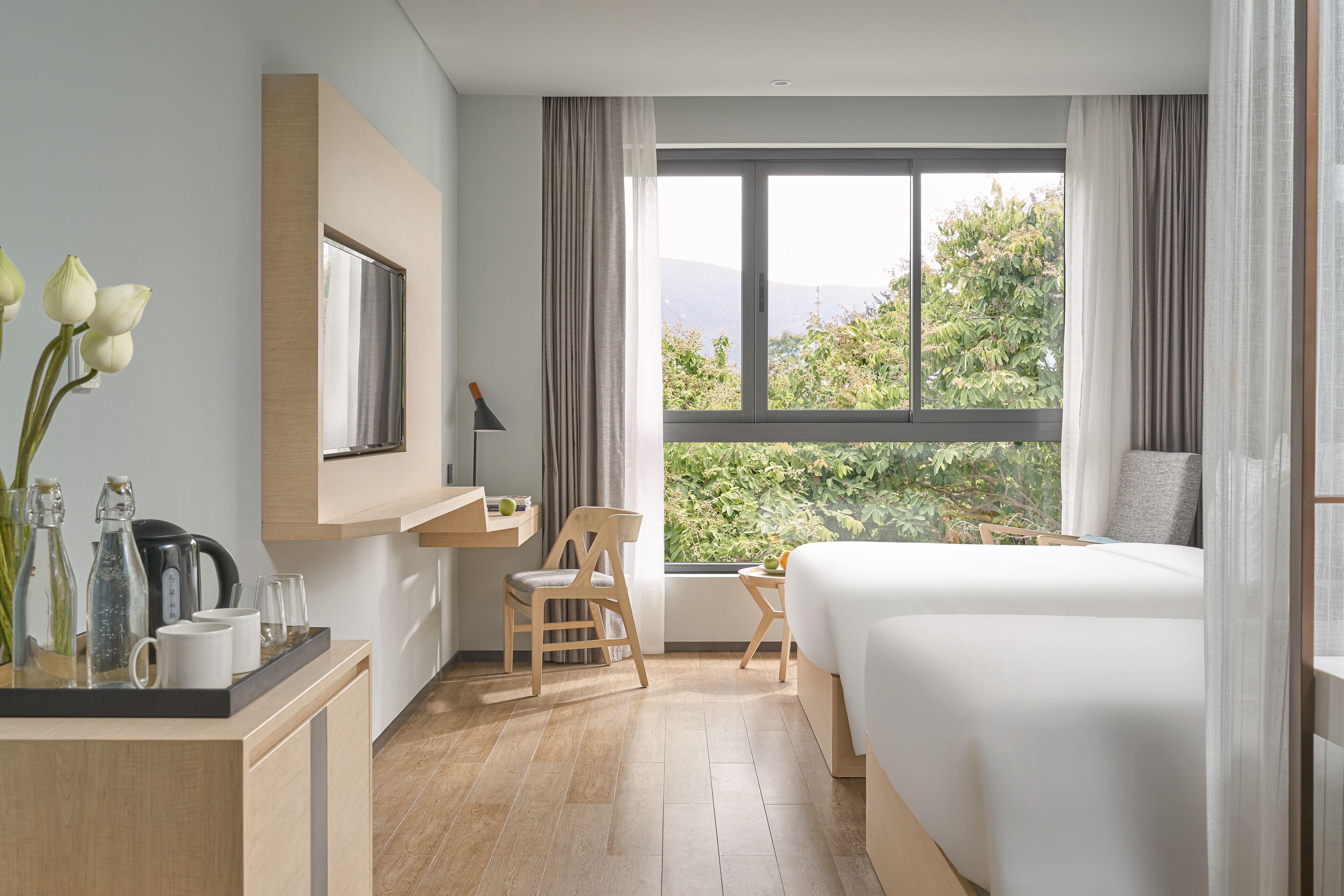 A modern hotel room with a large bed, a desk with a chair, a small table, and a view of greenery outside.
