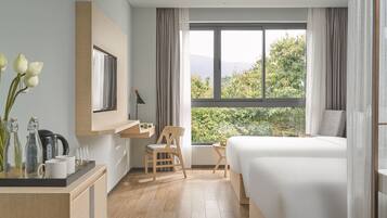 A modern hotel room with a large bed, a desk with a chair, a small table, and a view of greenery outside.
