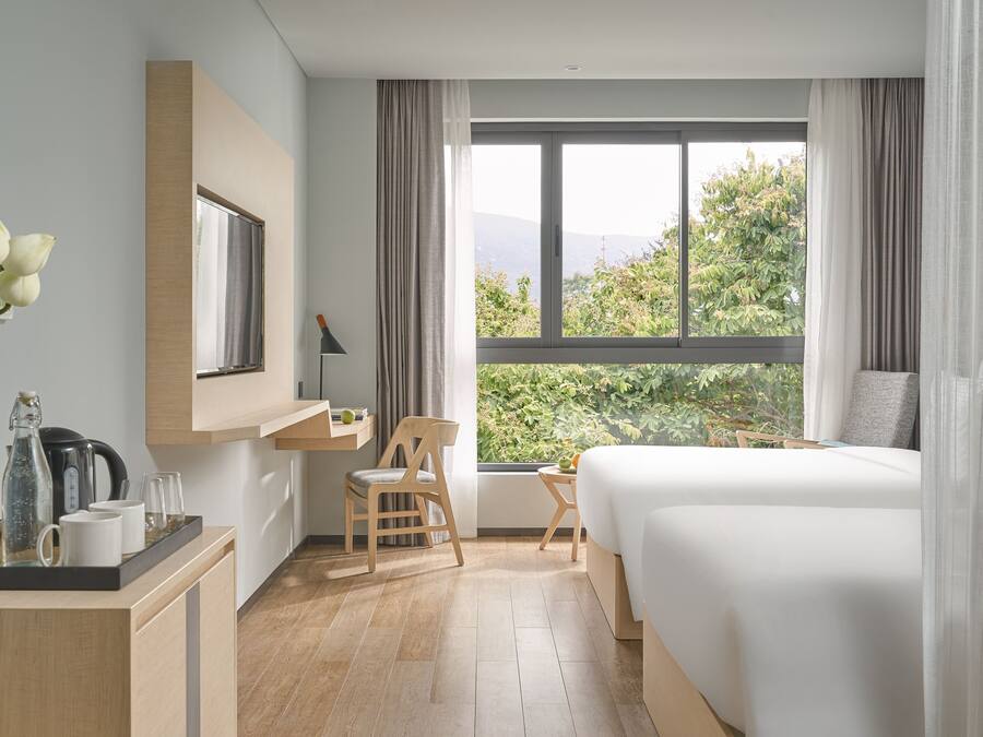 A modern hotel room with a large bed, a desk with a chair, a small table, and a view of greenery outside.