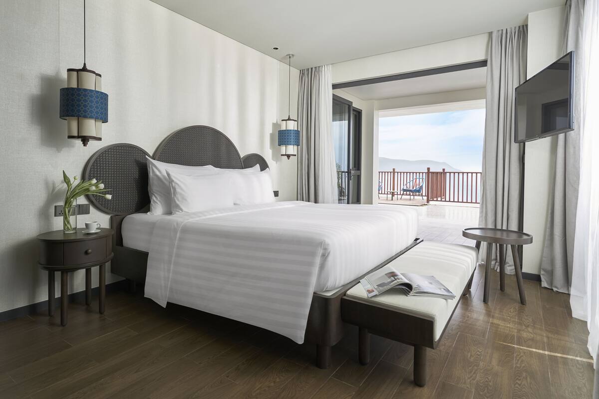 A hotel room with a large bed, bedside tables, a bench, a TV, and a balcony with a view.