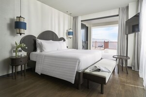 Habitación de hotel con una cama grande, mesitas de noche, un banco, un televisor y un balcón con vistas.