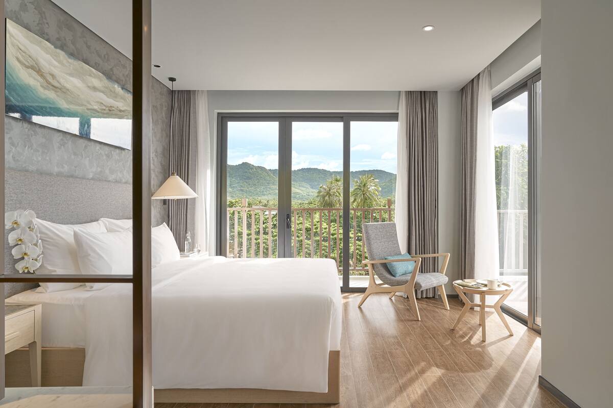 A hotel room with a large bed, a chair, a small table, and a view of mountains through large windows.