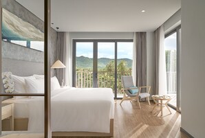 Camera d'albergo con un letto grande, una poltrona, un tavolino e vista sulle montagne attraverso ampie finestre.