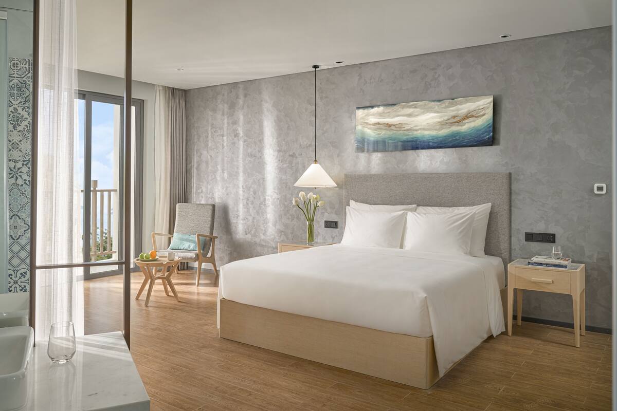 A modern hotel room with a large bed, a nightstand, a chair, a glass table, and a view of the outdoors.