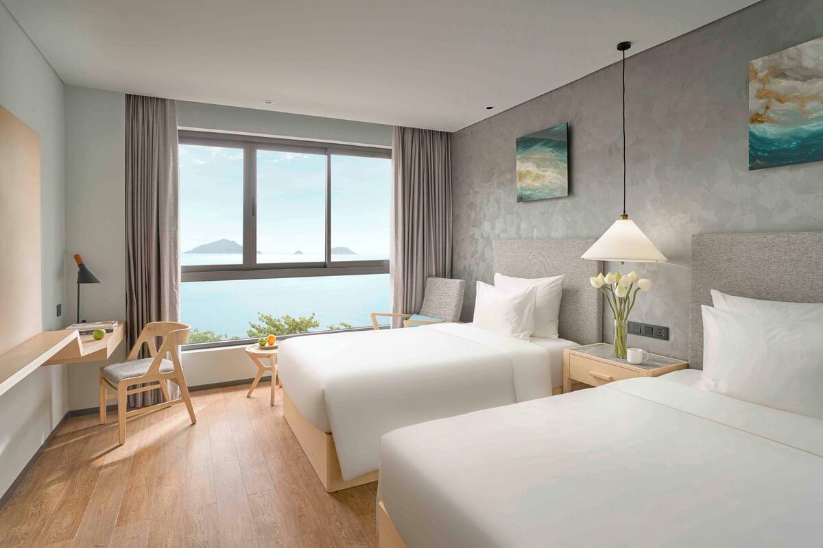 A hotel room with two beds, a desk, a chair, a nightstand, a lamp, and a view of the sea.