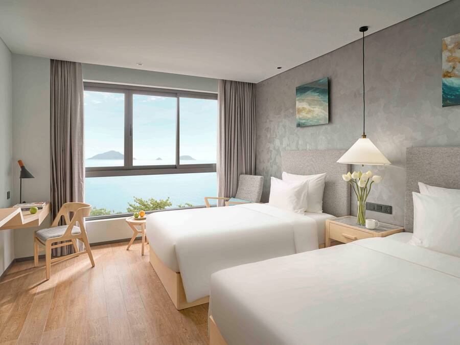 Habitación de hotel con dos camas, un escritorio, una silla, una mesita de noche, una lámpara y vistas al mar.