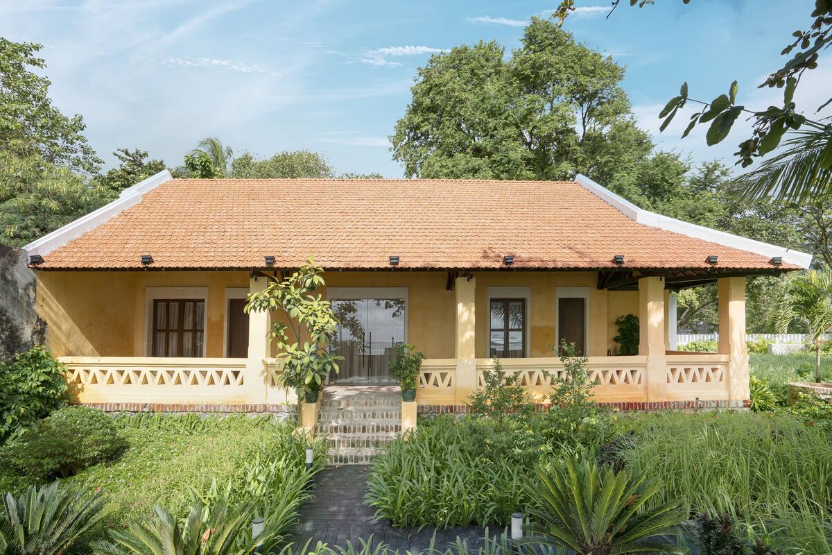 A yellow building with a tiled roof, a covered porch, and a garden with various plants.