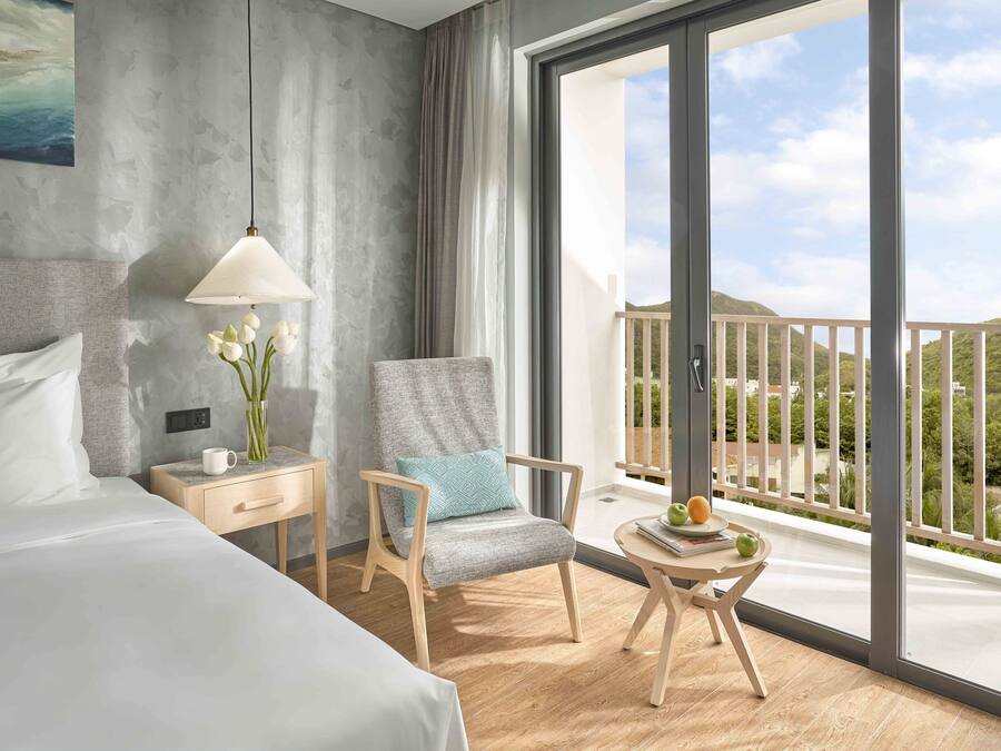 A hotel room with a bed, a chair, a nightstand, a lamp, and a view of the balcony.