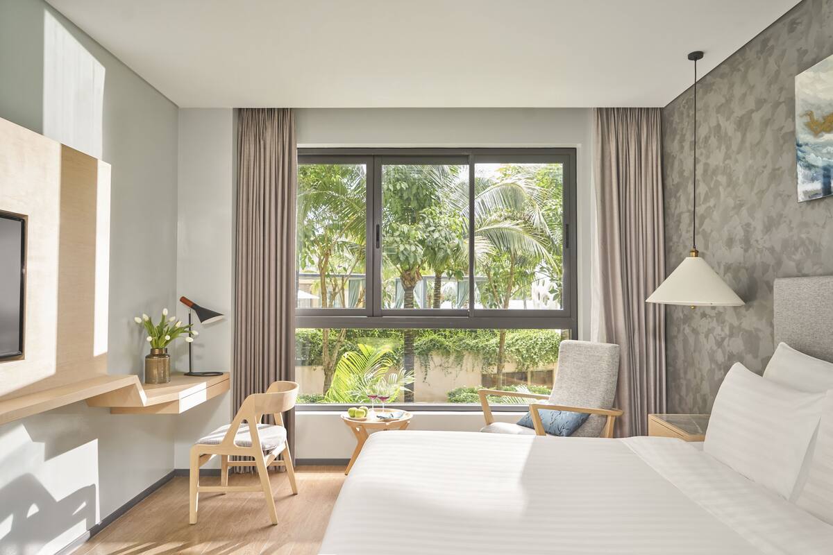 A hotel room with a large bed, a desk with a chair, a small table with drinks, and a view of greenery outside.