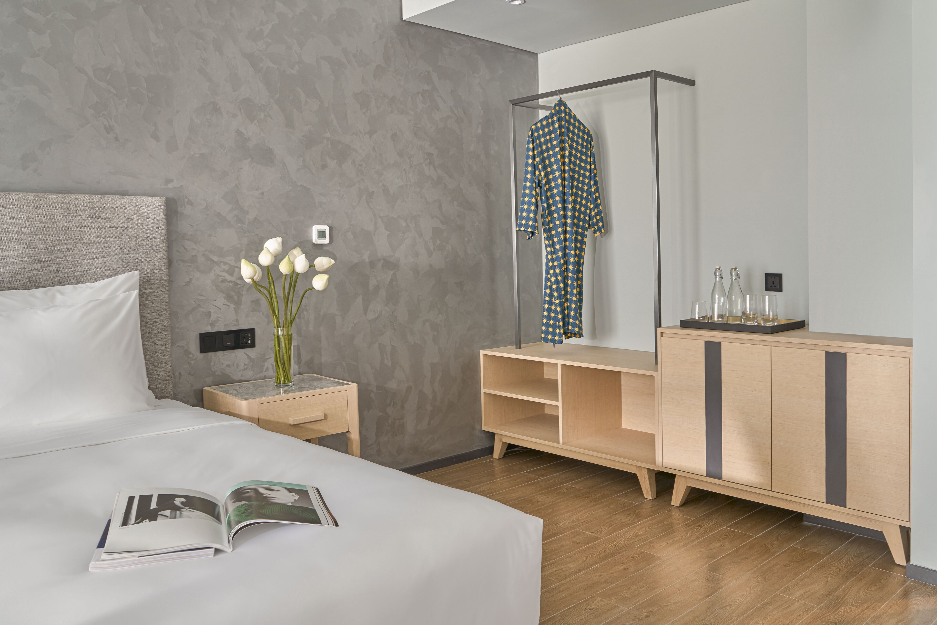 A modern hotel room with a large bed, a bedside table with a vase of flowers, a wooden dresser, and a mirror.