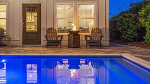 Un área junto a la piscina con una chimenea, dos sillas Adirondack y una puerta con panel de vidrio.