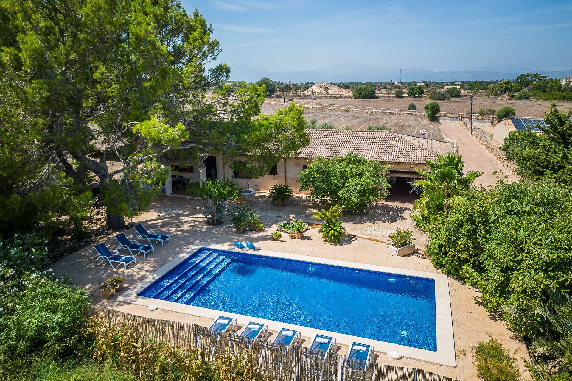 A swimming pool with lounge chairs, surrounded by greenery and a building with a tiled roof.