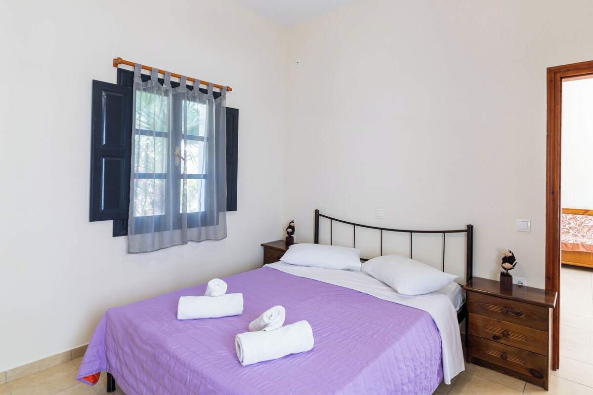 Una cama bien tendida con una colcha púrpura y almohadas blancas, una mesita de noche de madera con un adorno pequeño, y una ventana con cortinas.