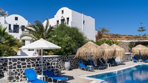 A pool area with blue lounge chairs, a stone wall, and a white building with a blue door. - Soulis Hotel (Santorini)