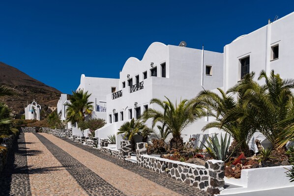 A white building with a stone pathway leading to it, surrounded by palm trees and rocks.