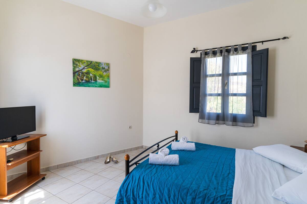 A bedroom with a bed, a TV on a wooden stand, a painting on the wall, and a window with curtains.