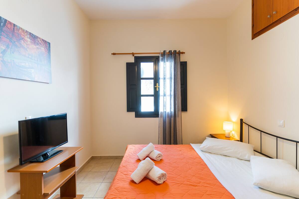 A hotel room with a bed, a TV on a wooden stand, a window with curtains, and a painting on the wall.