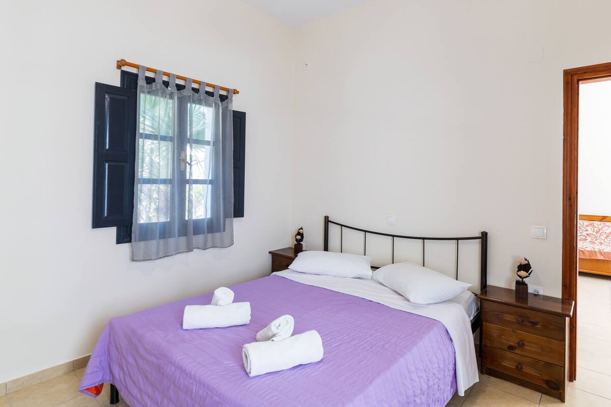 A neatly made bed with a purple bedspread and white pillows, a wooden nightstand with a small decorative item, and a window with curtains.