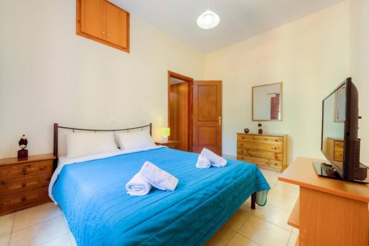A bedroom with a bed, wooden dresser, television, and a door leading to another room.