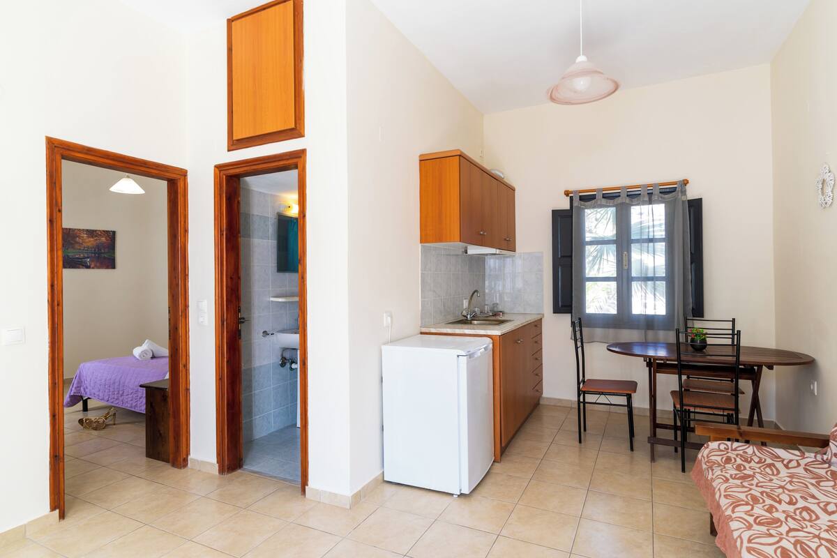 A compact kitchen with a refrigerator, wooden cabinets, a dining table with chairs, and a small window with curtains.