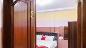 Une chambre d’hôtel avec une porte en bois portant le numéro 5, un lit avec une literie blanche et des touches de rouge, et un mur de couleur neutre en arrière-plan.