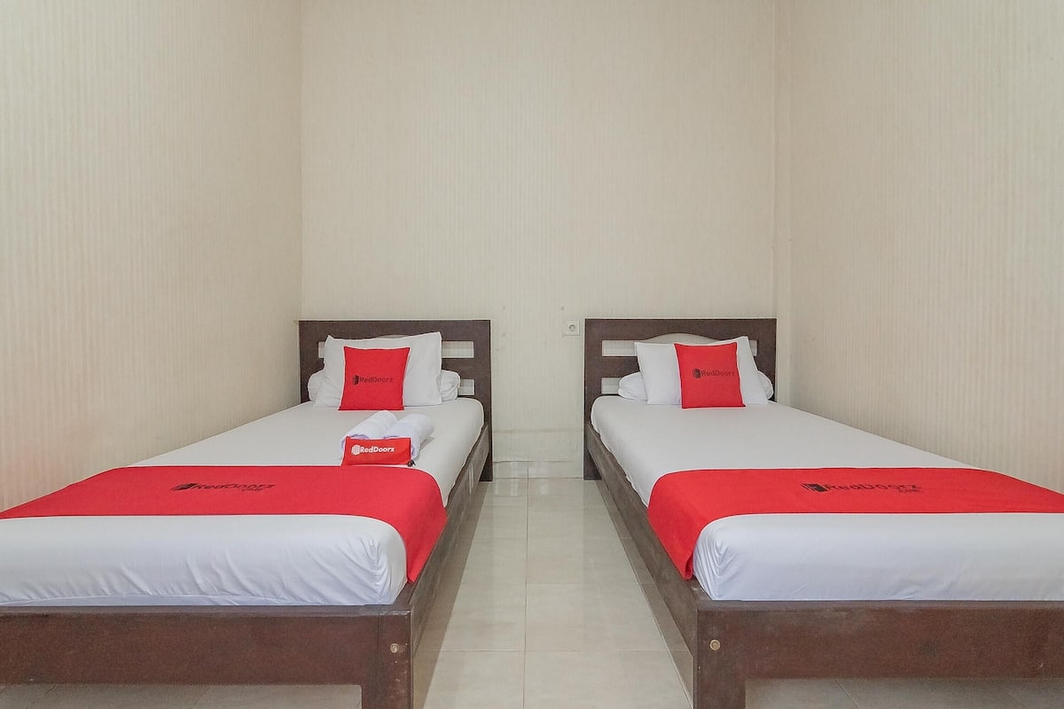 Two twin beds with white bedding and red accents, including pillows and a blanket, in a room with plain walls and a tiled floor.