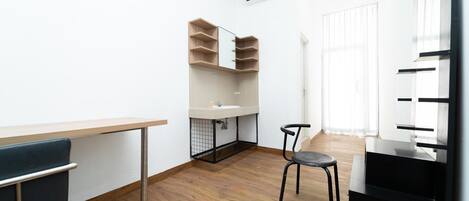A compact room with a wooden floor, a white sink, a mirror, a chair, and a black desk.