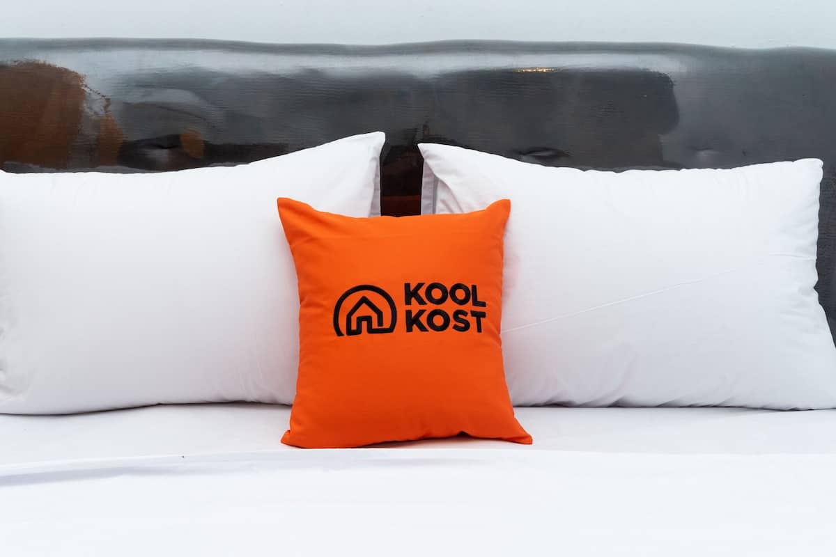Ein Bett mit weißen Kissen und einem orangefarbenen Kissen mit dem "Kool Kost"-Logo.