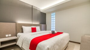 Una habitación de hotel moderna con una cama grande, una mesita de noche, una manta roja y un ventanal que deja entrar luz natural.