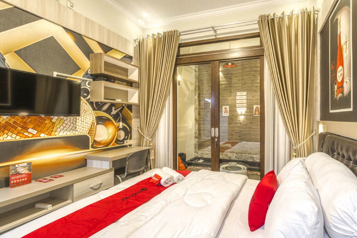 Habitación de hotel con cama, escritorio, silla, televisor y puerta de vidrio que da a un balcón.
