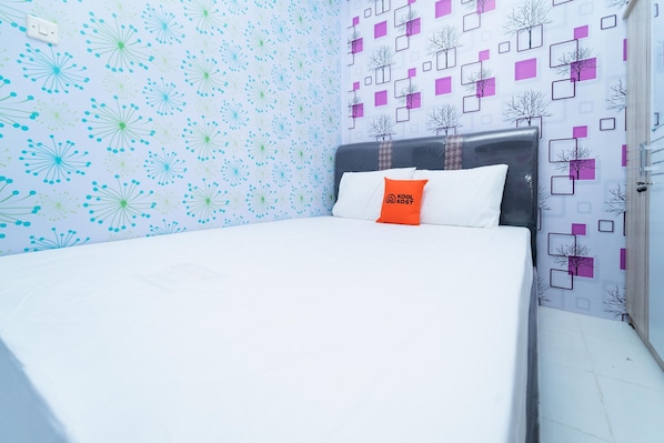 Un lit bien fait, avec un matelas blanc et un seul oreiller orange orné d’un logo. La pièce est décorée d’un papier peint à motifs bleus et blancs, et le mur adjacent présente un motif géométrique.