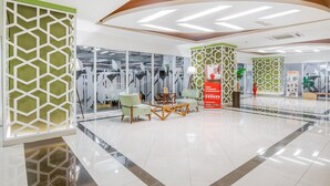 A modern lobby with geometric patterned walls, a seating area with light green chairs, and a central red information kiosk. - RedLiving Apartemen Grand Kamala Lagoon - Kita Pro Tower Barclay North (Bekasi)