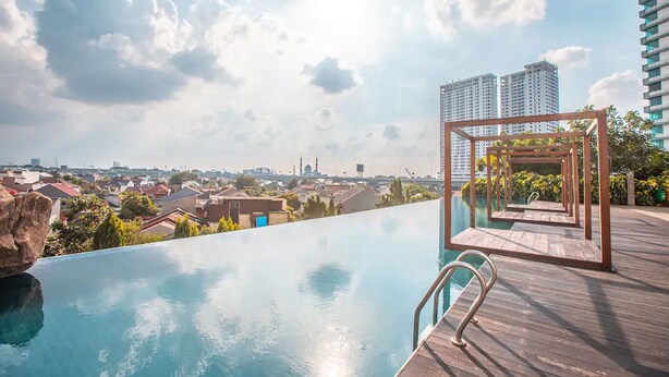 An infinity pool with a wooden deck and a view of a cityscape. - RedLiving Apartemen Grand Kamala Lagoon - Kita Pro Tower Barclay North (Bekasi)