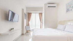 A modern hotel room with a large bed, a mounted TV, a small seating area, and a window with curtains. - RedLiving Apartemen Grand Kamala Lagoon - Kita Pro Tower Barclay North (Bekasi)