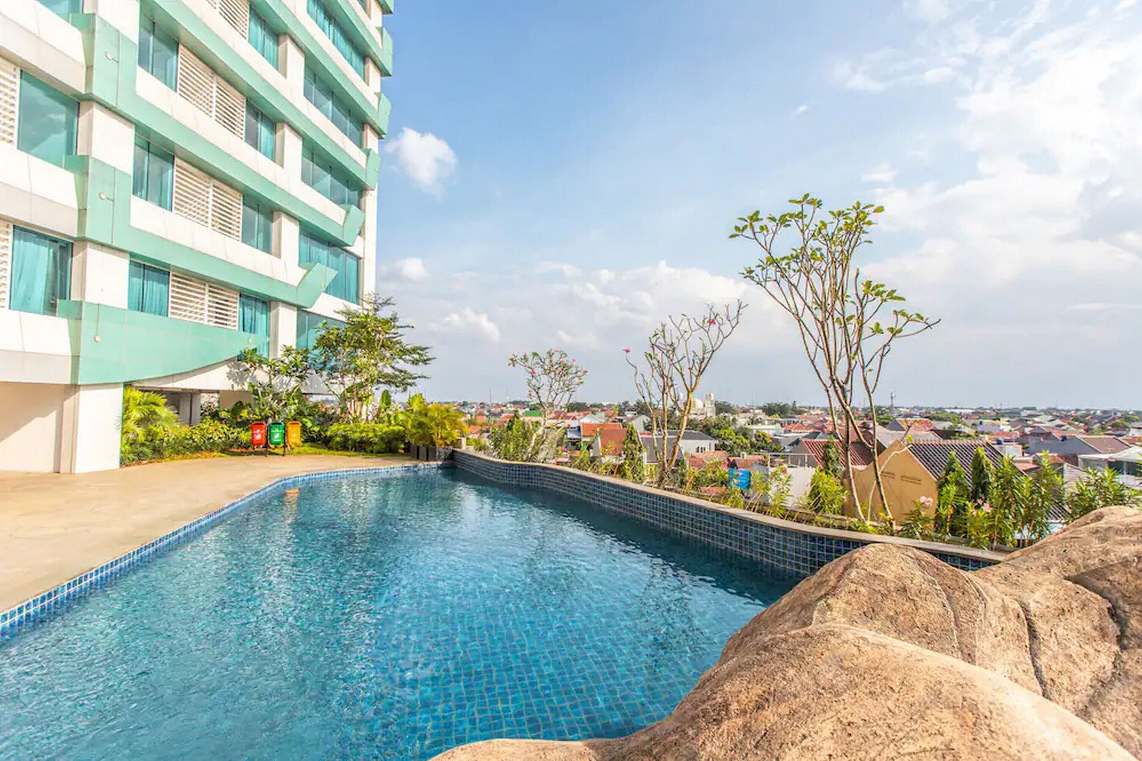 A swimming pool with a tiled edge, surrounded by rocks and greenery, with a view of a multi-story building and a residential area in the background.