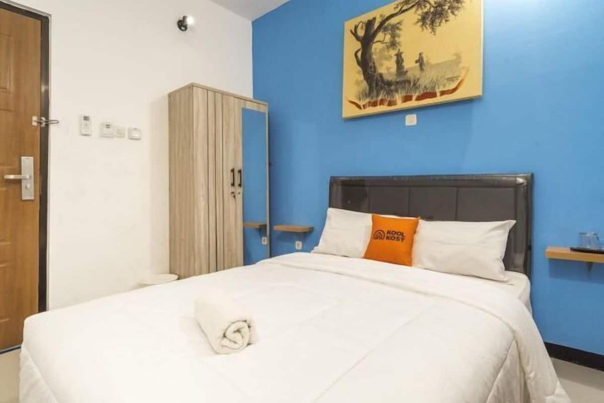 A hotel room with a bed, a wardrobe, a painting on the wall, and a small shelf.