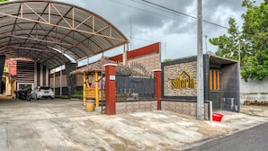 A covered parking area with a gated entrance, a small building with a stone facade, and a red bucket on the ground. - RedDoorz Syariah near Kebon Rojo Park Blitar (Blitar)