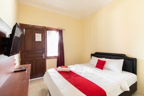 A hotel room with a bed, a TV on a wooden desk, a door, and a window with curtains. - RedDoorz @ Raya Janti Yogyakarta (Baturetno)