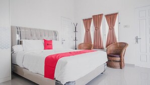 A modern hotel room with a large bed, a red accent pillow, two wicker chairs, and a coat rack. - RedDoorz Syariah near Arafah Hospital Jambi (Jambi)