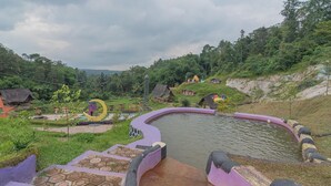 Una piscina de borde curvo, rodeada por una barandilla morada y negra, ubicada en un paisaje verde y exuberante con colinas y chozas tradicionales al fondo.