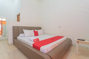 Un lit bien fait, avec une couverture rouge et des oreillers, une table de chevet en bois avec une télécommande, et une fenêtre avec des rideaux.