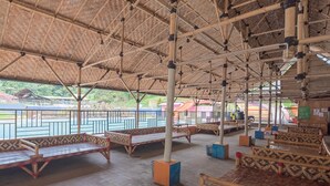 Un pabellón descubierto con vigas de bambú y suelo de baldosas, con bancos decorados y vista a un parque infantil.