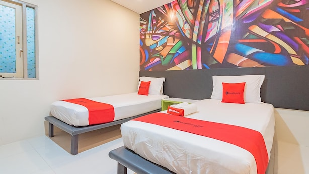 Une chambre d’hôtel avec deux lits, chacun agrémenté de draps blancs et de touches de couleur rouge, une fresque murale abstraite et colorée, et une fenêtre avec du verre dépoli.