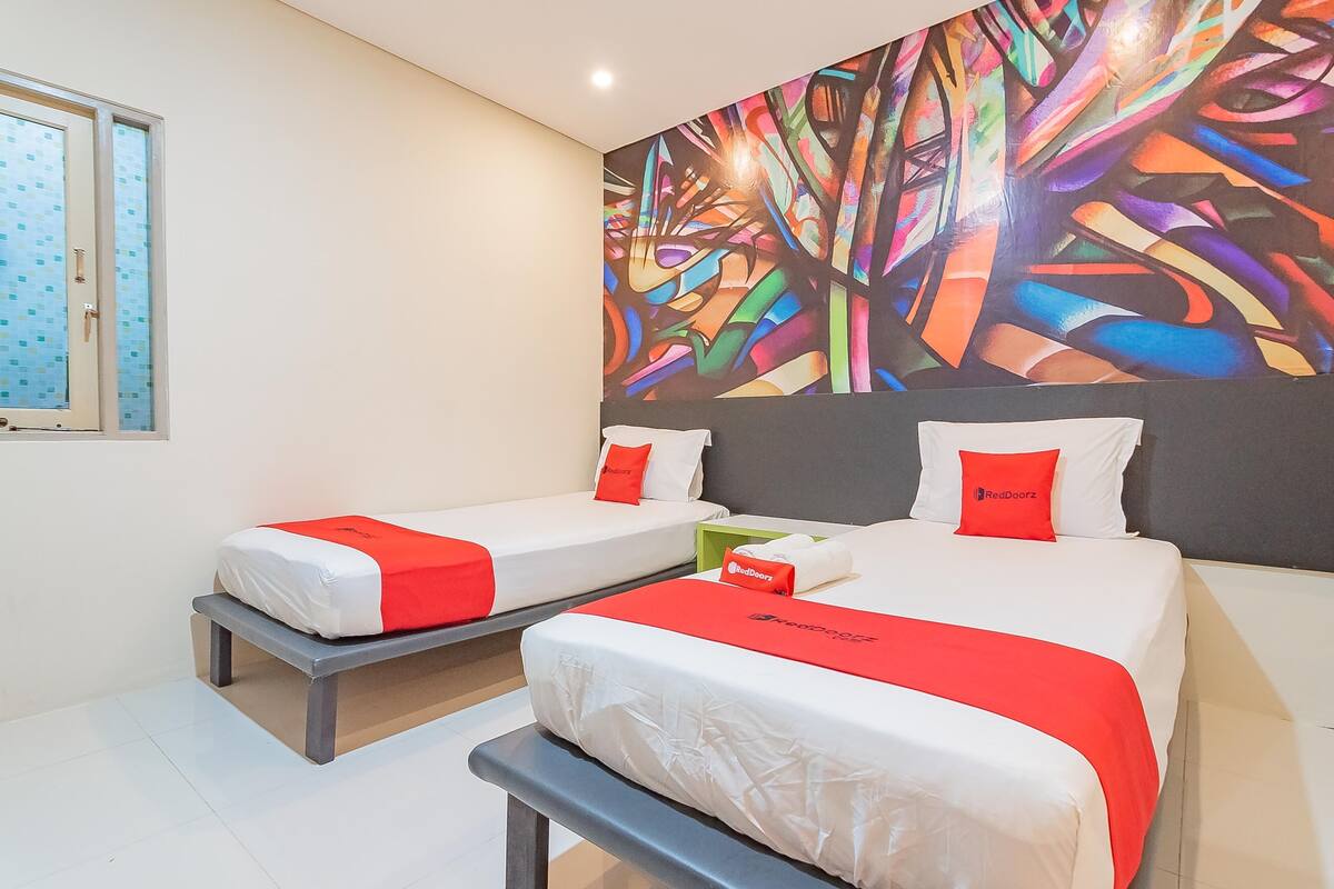 A hotel room with two beds, each with white bedding and red accents, a colorful abstract mural on the wall, and a window with frosted glass.