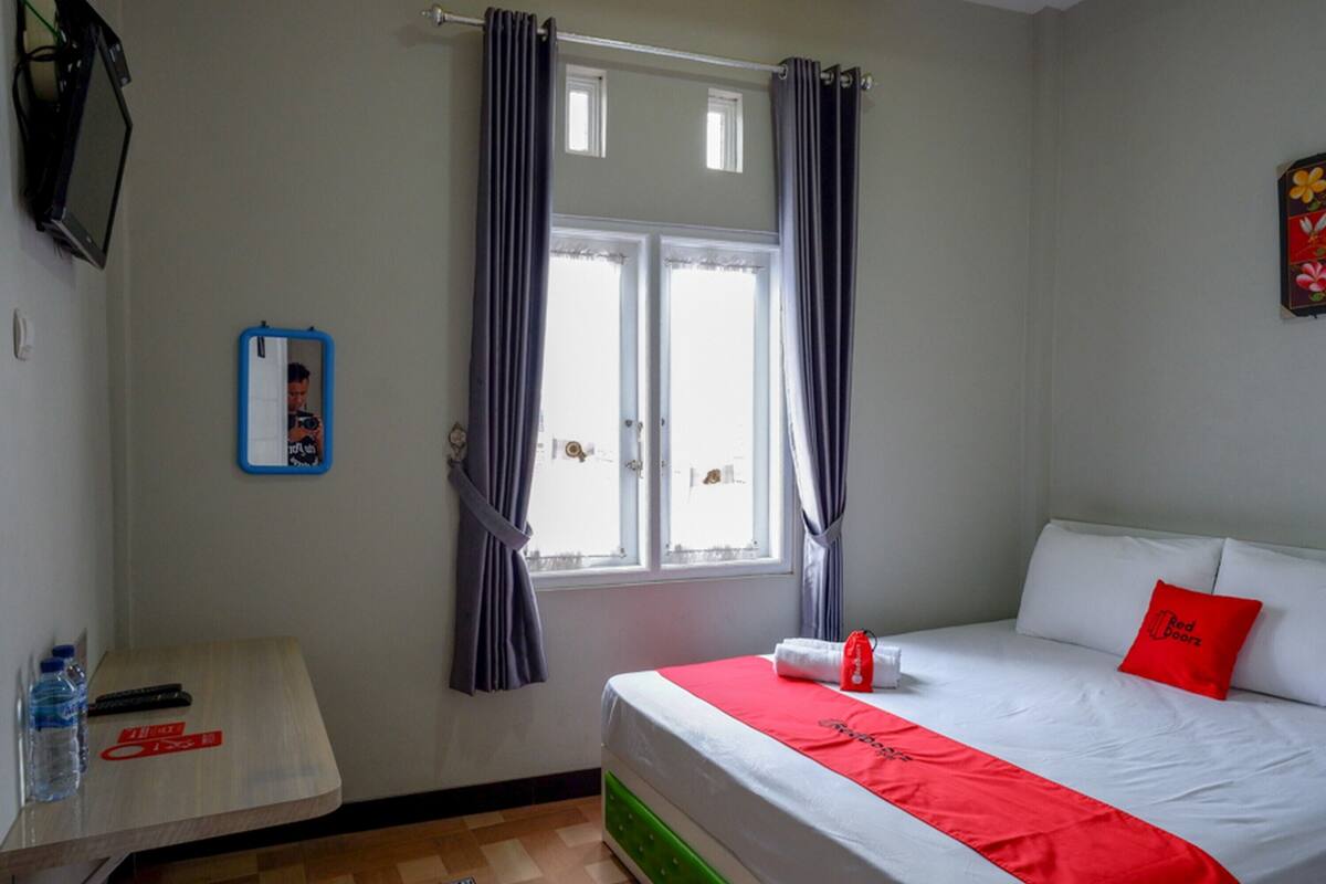 Habitación de hotel con cama, una mesita con una botella de agua y un control remoto, una ventana con cortinas y un televisor montado en la pared.