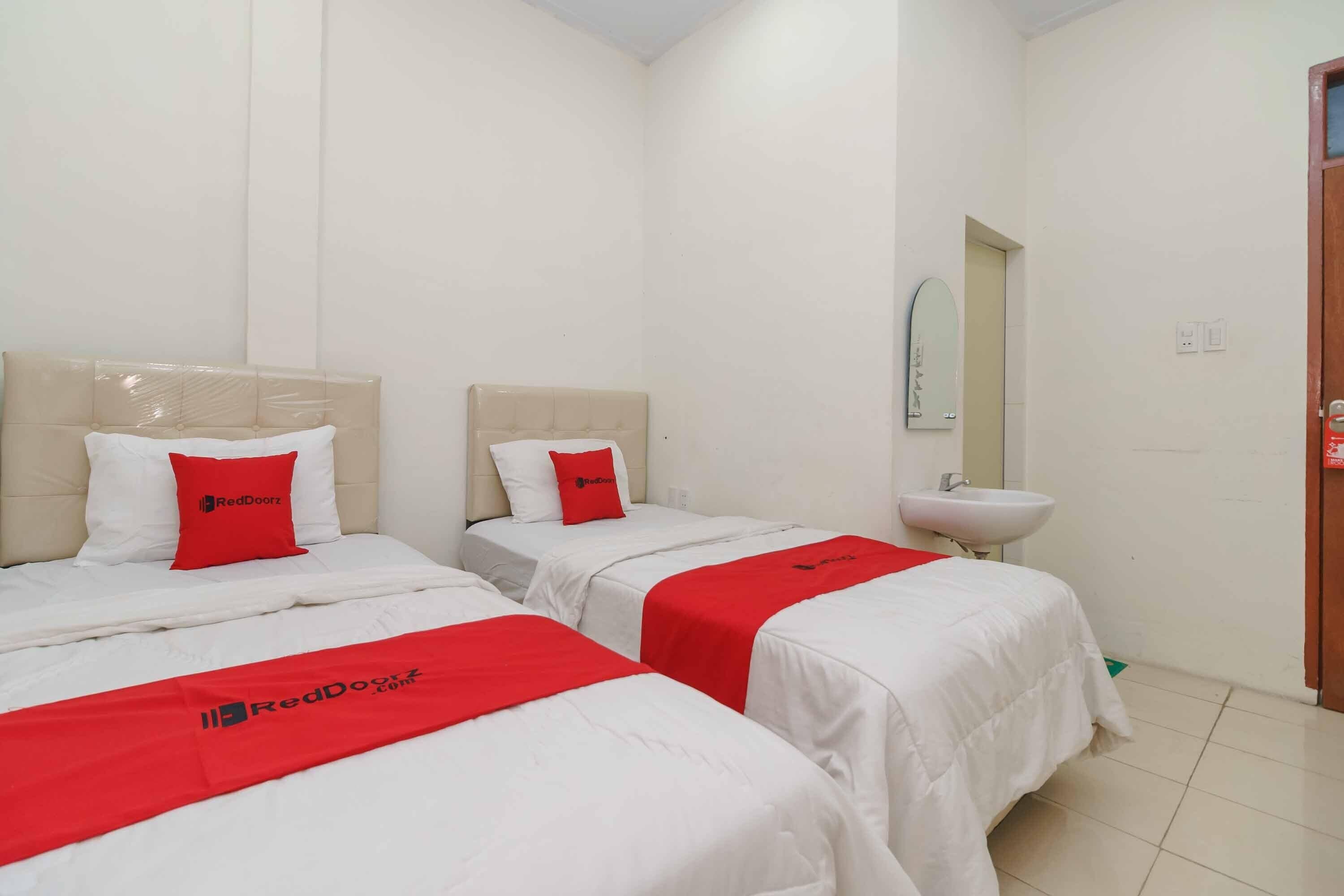A hotel room with two beds, each with a white bedspread and a red blanket. There is a white sink and a mirror on the wall.