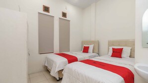 A hotel room with two beds, each with a red bedspread and white bedding. There are two windows with beige curtains, a white sink, and a door.