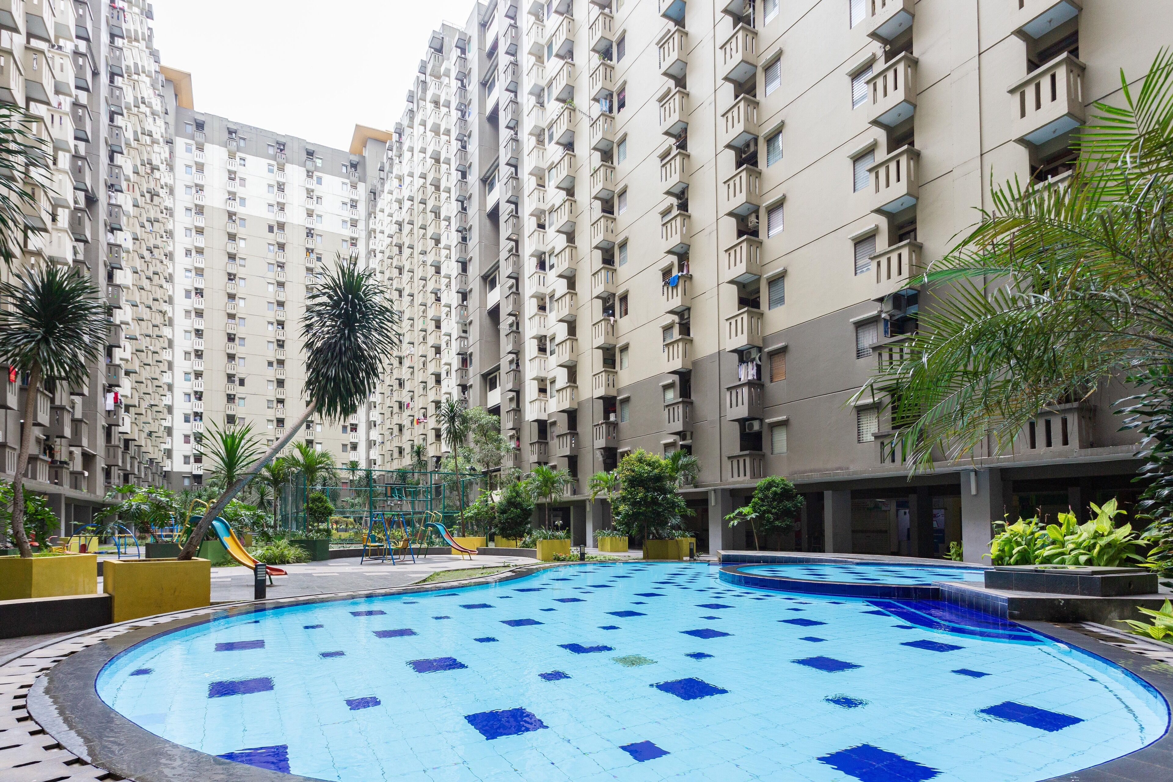 Piscina cercada por edifícios residenciais altos, com um parquinho e vegetação em primeiro plano.